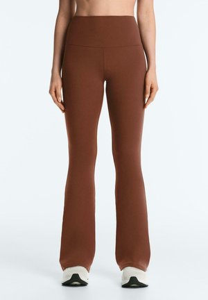 Kvinde iført højtaljede, brune, elastiske bukser med vidde forneden, stående mod en ensfarvet baggrund, kombineret med hvide sneakers.