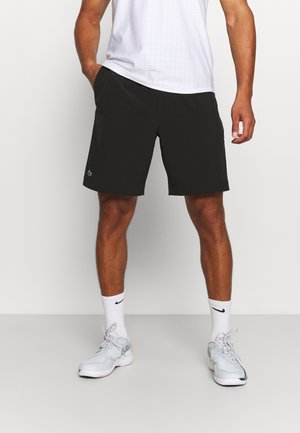 Mężczyzna stojący w czarnych spodenkach, białych skarpetkach, białych butach sportowych Nike oraz białej koszulce sportowej, z rękami luźno opuszczonymi, jedną w kieszeni.