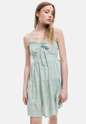 Helles grünes, gestuftes Kleid aus strukturiertem Stoff mit Lochstickerei. Verfügt über dünne Träger und einen geraffen Rüschenausschnitt mit einer Schleife.