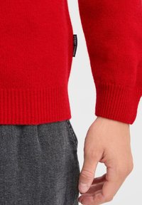 Czerwony sweter z dzianiny z prążkowanymi mankietami, z metką po boku. Dodatkowo ciemnoszare spodnie o gładkiej fakturze tkaniny.