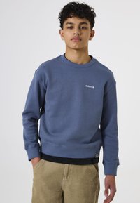 Sudadera azul hecha de un tejido suave con cuello redondo y puños acanalados. Logo "GARCIA" en blanco en la parte frontal. Llevada sobre pantalones de pana.