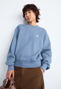 Light blue sweatshirt with a round neckline and ribbed cuffs; paired with brown pants and a pink handbag. Notable small logo on chest.
