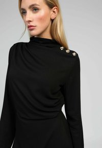 Femme aux longs cheveux blonds portant un haut noir à manches longues avec trois boutons décoratifs dorés sur l'épaule.