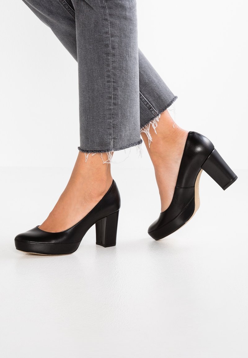 Escarpins noirs en cuir à talons hauts avec un bout rond, une texture lisse et un talon carré épais, associés à un jean gris à ourlet effiloché.