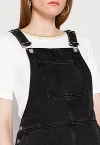 Black denim overalls featuring adjustable silver hardware and a front pocket, worn over a white shirt with subtle colored stripes.