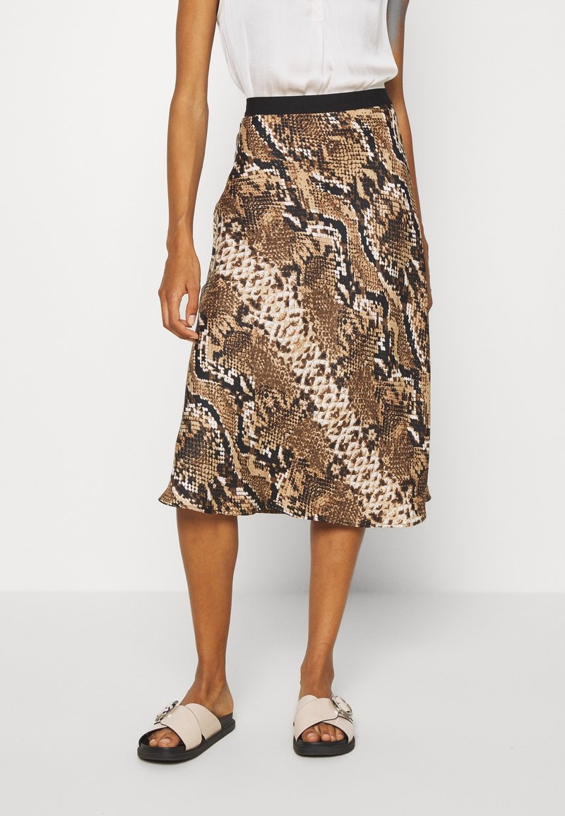 Femme portant une jupe midi imprimé serpent marron et noir, un chemisier blanc sans manches et des sandales beige à enfiler avec des boucles.