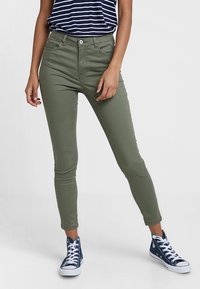 Jeans skinny vert olive à taille haute, avec des poches avant et une fermeture à bouton, associés à une chemise rayée bleu marine et des baskets bleues.