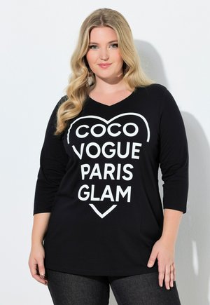 Vaaleahiuksinen nainen, jonka hiukset ovat laineikkaat, pukeutuneena mustaan toppiin, jossa on kolmen neljänneksen hihat ja valkoinen teksti "COCO VOGUE PARIS GLAM" sydämen muotoisessa kehyksessä.