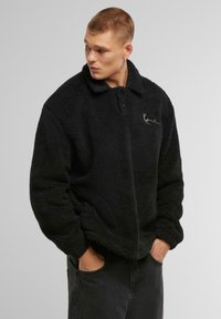 Pullover in pile nero con una consistenza morbida, collo a camicia e apertura con bottoni. Presenta un logo ricamato sul lato sinistro del petto.