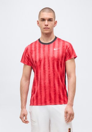 Junger Mann mit kurzen Haaren trägt ein rotes Nike-Sport-T-Shirt und weiße Shorts, steht vor einem schlichten hellen Hintergrund.