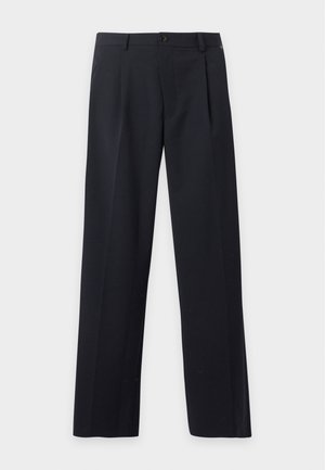 Homme portant des lunettes de soleil, une veste sombre avec de grandes poches, une chemise blanche, un pantalon sombre et des chaussures noires, debout devant un fond blanc uni.