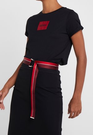 Persona con camiseta negra ajustada con logotipo rojo de HUGO, falda negra y un cinturón largo rojo con rayas blancas colgando.