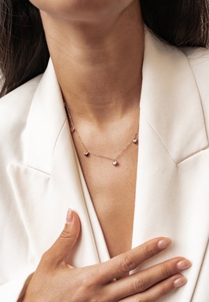 Woman wearing a white blazer and delicate gold necklace with small heart-shaped charms, hand resting on her chest.