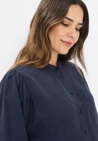 Marineblå bluse med høj krave, knaplukning foran og puffede ærmer. Stoffet har en glat tekstur. Har diskrete stikninger.