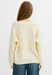 Suéter de punto color crema con un ajuste suelto y textura acanalada, con hombros caídos y dobladillo trasero ligeramente más largo, combinado con jeans azules.