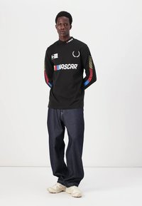Svart langermet skjorte med hvit "NASCAR" logo og fargerike striper på ermene, kombinert med løse mørke denimjeans og lyse sneakers.
