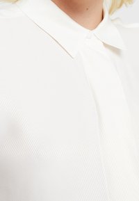 Bílá blůza s límečkem, která má jemné vertikální pruhy. Materiál má hladkou texturu, což vytváří čistý a sofistikovaný vzhled.