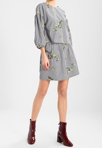 Schwarz-weiß kariertes Kleid mit gelben Blumenmustern, hat puffige Ärmel, einen runden Halsausschnitt und wird mit bordeauxfarbenen Knöchelstiefeln kombiniert.