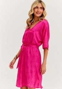 Ružové šaty s výstrihom do V, krátkymi rukávmi a opaskom v páse. Hladká látka má jemný vzor a siaha po polovicu lýtok.