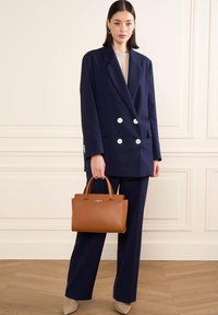 Blazer doppiopetto blu navy con bottoni argentati, pantaloni abbinati e una borsa in pelle marrone. Tessuti morbidi e vestibilità strutturata.