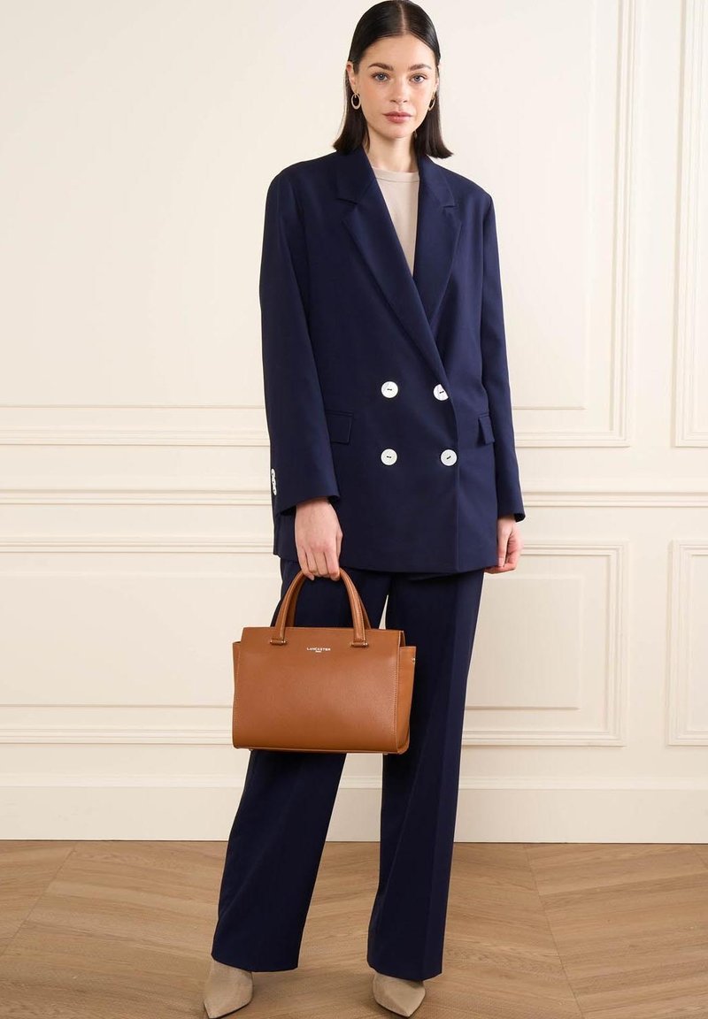 Blazer doppiopetto blu navy con bottoni argentati, pantaloni abbinati e una borsa in pelle marrone. Tessuti morbidi e vestibilità strutturata.