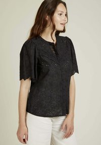 Blouse noire à manches courtes, avec un design floral texturé et une fermeture à boutons au cou, fabriquée en tissu léger.