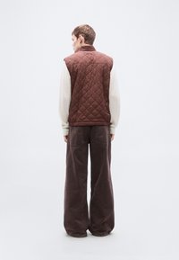 Καφέ καπιτονέ γιλέκο με ψηλό γιακά, φορεμένο πάνω από ανοιχτόχρωμο πουκάμισο με μακριά μανίκια, συνδυασμένο με φαρδιά, σκουρόχρωμα παντελόνια.