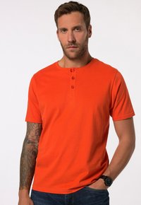 Oranžové tričko s krátkým rukávem a kulat�ým výstřihem s knopkovou légou na čtyři knoflíky. Model ho nosí s tmavými džíny a černými náramkovými hodinkami.