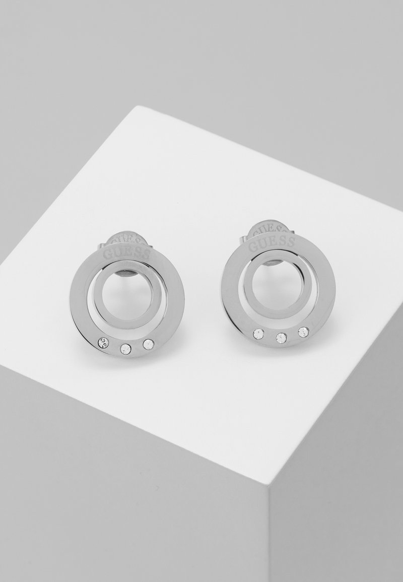 Boucles d'oreilles clous circulaires en argent avec trois petits cristaux et le nom de la marque Guess gravé sur chacune, exposées sur une surface blanche.