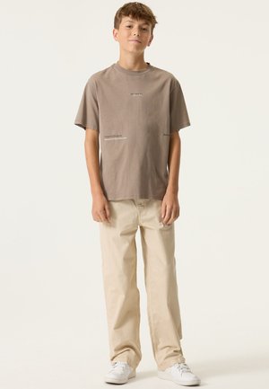 Bruine katoenen T-shirt met tekstprints op de voorkant, gecombineerd met crème-kleurige, losvallende broek en witte sneakers.