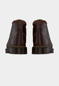 Ζευγάρι από σκούρα καφέ δερμάτινα μποτάκια φαίνονται από πίσω, με χοντρές μαύρες σόλες και ορατές ραφές σε λευκό φόντο.