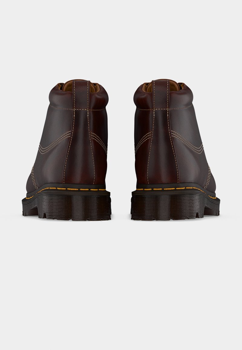 Een paar donkerbruine leren laarzen gezien van achteren, met dikke zwarte zolen en zichtbare stiksels, op een witte achtergrond.