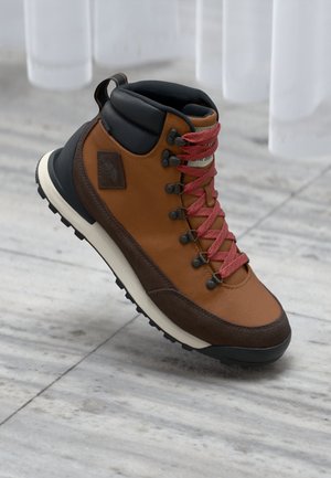 Brown hiking boot with black padded collar, red laces, and rugged black sole on marble floor background.