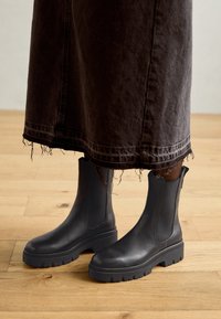 Botas de tobillo de cuero negro con paneles elásticos en los laterales, suelas de goma gruesa y una textura suave, combinadas con una falda de mezclilla oscura y desgastada.
