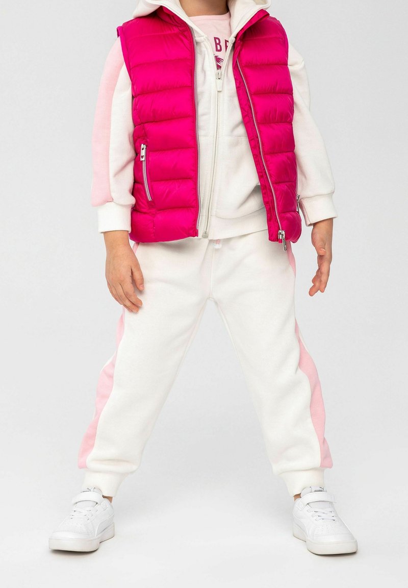 Enfant portant un survêtement blanc et rose avec des baskets blanches et un gilet matelassé sans manches rose vif.