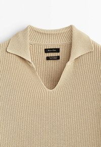 Beżowy sweter dzianinowy z dekoltem w kształcie litery V, prążkowanym kołnierzem i teksturowanym materiałem. Posiada dwie wewnętrzne etykiety w czarnym kolorze, z informacjami o marce i pielęgnacji.