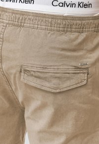 Beige puuvillashortsit, joissa on yksi takatasku ommellulla yksityiskohdalla ja Calvin Klein -logo näkyvissä ylhäällä vyötäröllä.