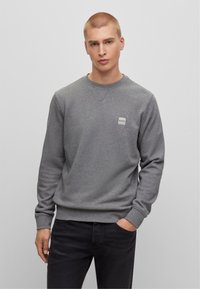 Giovane con capelli biondi corti, che indossa una felpa grigia BOSS e jeans neri, in piedi con una mano in tasca su uno sfondo grigio uniforme.