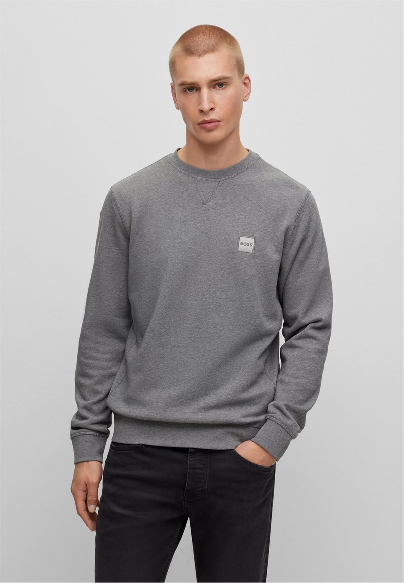 Giovane con capelli biondi corti, che indossa una felpa grigia BOSS e jeans neri, in piedi con una mano in tasca su uno sfondo grigio uniforme.