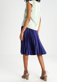 Blusa verde chiaro con maniche arricciate, abbinata a una gonna plissettata blu scuro. Tacchi beige con cinturino alla caviglia completano l'outfit.