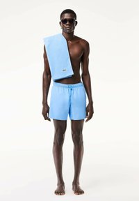 Niebieskie kąpielówki z elastycznym pasem, połączone z pasującym jasnoniebieskim ręcznikiem zwisającym z ramienia. Okulary przeciwsłoneczne dopełniają stylizację.