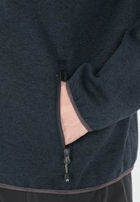 Tummanharmaa vetoketjullinen takki, jossa on teksturoitu kangas, sivutasku ja tummanruskeat korostukset hihansuissa ja helmais. 