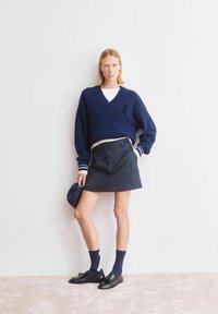 Marineblauer Strickpullover mit V-Ausschnitt und cremefarben gestreiften Ärmeln, kombiniert mit einem marineblauen Nadelstreifenrock und dunklen Schuhen, mit einem schwarzen Banddetail.