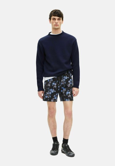 Marineblauer Pullover mit strukturierter Oberfläche, kombiniert mit schwarzen Shorts mit Blumenmuster in Blau und Weiß. Schwarze Sportschuhe und Socken runden den Look ab.