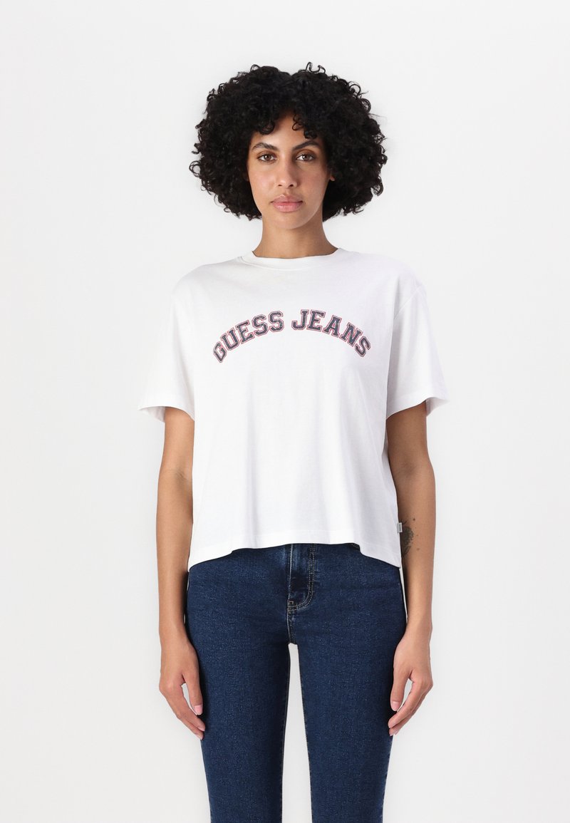 Balta apkirpta maroška su "GUESS JEANS" ryškiu, spalvotu tekstu. Savybės: trumpi rankoviai ir apvalus kaklas, derinama su tamsiai mėlynomis džinsomis.