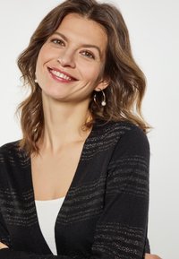 Cardigan negru cu model de dungi metalice, tricotat texturat și design în formă de V. Purtați peste un top alb, cu cercei minimalisti tip cerc.