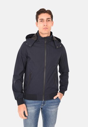 Giovane uomo con giacca blu navy con cerniera e cappuccio e jeans blu, in piedi con una mano in tasca contro uno sfondo chiaro neutro.