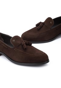 Mocasines de ante marrón con detalles de borlas, puntera redondeada, costuras en contraste y una suela de goma negra. Dos costuras decorativas laterales.