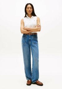 Witte mouwloze polo, high-waisted blauwe denim wijd-hangende jeans, bruine leren riem en bruine instappers met een zilveren accent.