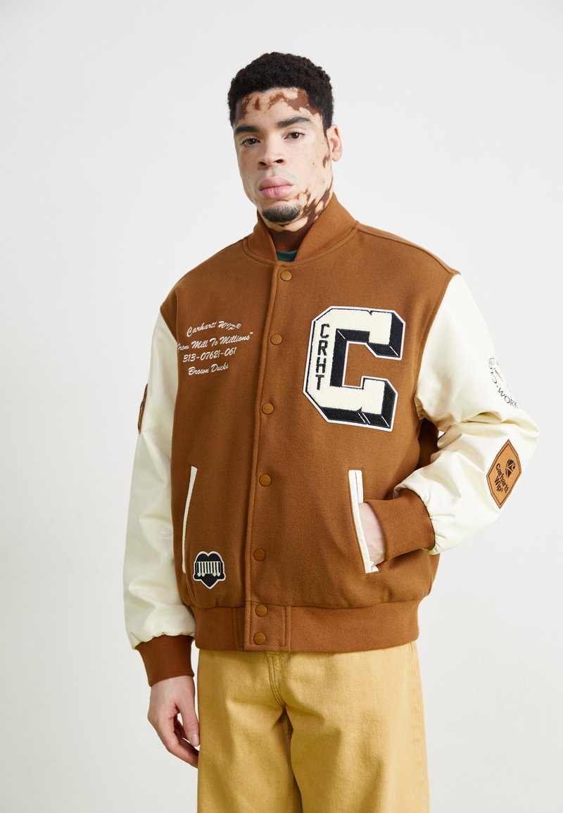 Smeđi sveučilišni kaput s kremastim rukavima, s velikim slovom "C," izvezenim detaljima, gumbima na preklapanje i dva prednja džepa.